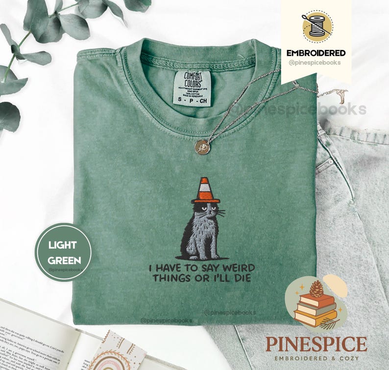 May include: Light green t-shirt with a graphic of a cat wearing a traffic cone hat. The text "I HAVE TO SAY WEIRD THINGS OR I'LL DIE" is below the cat. The shirt has a crew neck and short sleeves.