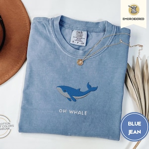May include: Pale blue t-shirt with an embroidered blue whale design and the text "OH WHALE". The shirt is made of 100% cotton and is pre-shrunk. A gold necklace and a brown hat are also in the image.