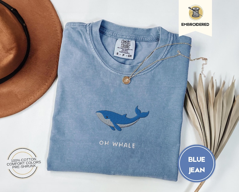 May include: Pale blue t-shirt with an embroidered blue whale design and the text "OH WHALE". The shirt is made of 100% cotton and is pre-shrunk. A gold necklace and a brown hat are also in the image.