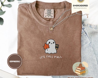 May include: Espresso-colored t-shirt with an embroidered ghost holding a maple leaf and a coffee cup. The text "IT'S FALL Y'ALL" is below the design. The shirt has a crew neck and is labeled "COMFORT COLORS". A silver necklace is draped across the shirt.