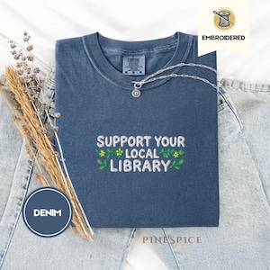 Op de afbeelding: Een denim blauw t-shirt met de geborduurde tekst "SUPPORT YOUR LOCAL LIBRARY" in wit, groen en geel. Het shirt is opgevouwen en wordt gepresenteerd met een zilveren ketting en gedroogde bloemen.