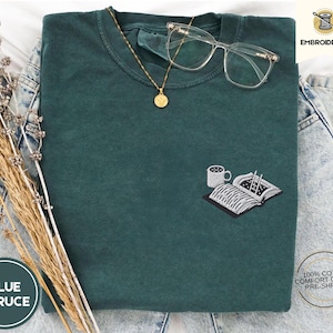 May include: A dark green t-shirt with a white embroidered design of a book, a cup of coffee, and a pair of chopsticks. The text "EMBROIDERED" is in the top right corner of the image. The text "BLUE SPRUCE" is in the bottom left corner of the image.