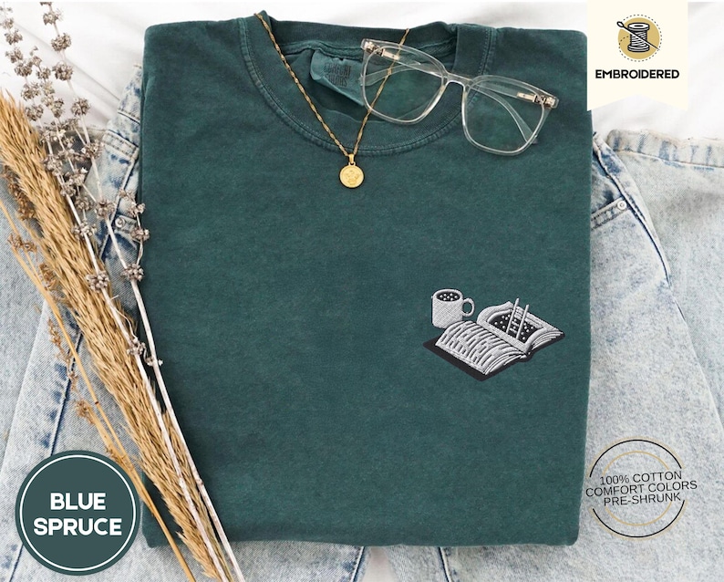 May include: A dark green t-shirt with a white embroidered design of a book, a cup of coffee, and a pair of chopsticks. The text "EMBROIDERED" is in the top right corner of the image. The text "BLUE SPRUCE" is in the bottom left corner of the image.
