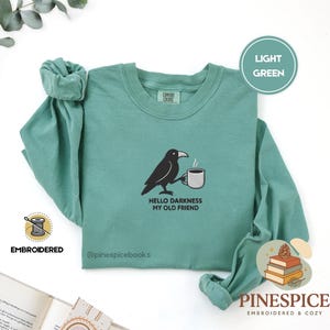 Op de afbeelding: Een lichtgroen sweatshirt met lange mouwen en een ronde hals, voorzien van een geborduurde zwarte kraai die een witte koffiemok vasthoudt. Onder de kraai staat de tekst "HELLO DARKNESS MY OLD FRIEND" in zwart borduurwerk.