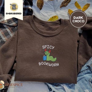Puede incluir: Sudadera marr&oacute;n chocolate oscuro con el texto bordado "SPICY BOOKWORM" y un dise&ntilde;o de gusano de libro de dibujos animados. El gusano de libro es verde con gafas y peque&ntilde;os cuernos rojos. Estilo c&oacute;modo e informal.