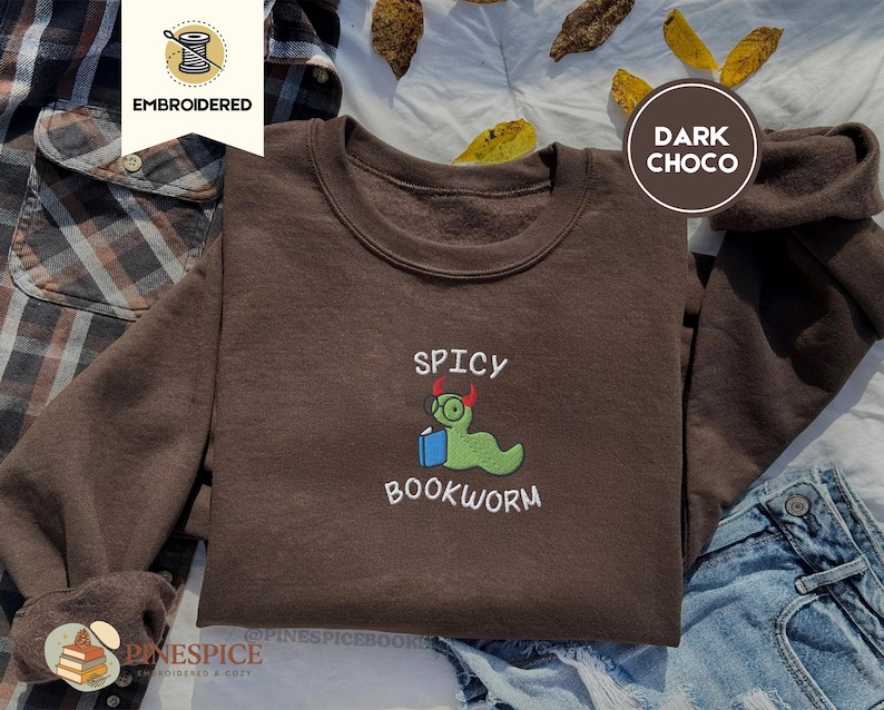 Puede incluir: Sudadera marr&oacute;n chocolate oscuro con el texto bordado "SPICY BOOKWORM" y un dise&ntilde;o de gusano de libro de dibujos animados. El gusano de libro es verde con gafas y peque&ntilde;os cuernos rojos. Estilo c&oacute;modo e informal.