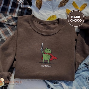May include: Dark chocolate-colored sweatshirt featuring a cartoon frog in a red cape, holding a sword. The frog is green with a red bow tie. The word "Huzzah" is printed below the frog. The sweatshirt is displayed with a plaid shirt and denim jeans.