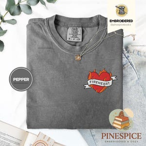May include: A grey Comfort Colors t-shirt with a 'Fireheart' embroidered design. The design features a red heart with flames and a white banner. A silver necklace with a pendant is also visible.