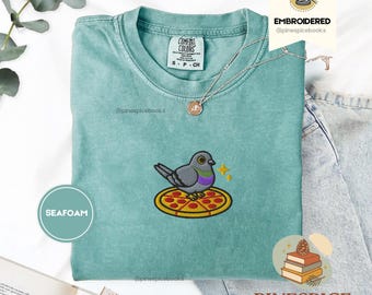 May include: Seafoam green t-shirt with an embroidered design of a gray pigeon perched on a pizza. The shirt has a crew neck and short sleeves. The text "COMFORT COLORS" is printed on the upper left side of the shirt.