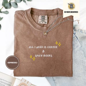 Op de afbeelding: Espresso-kleurig T-shirt met geborduurde tekst "All I Need Is Coffee & Spicy Books." Het shirt heeft een ronde hals en korte mouwen. Een zilveren ketting met een hanger is over het shirt gedrapeerd. Het shirt is op een wit oppervlak gevouwen.