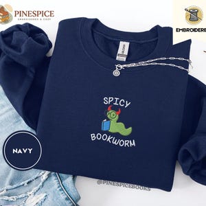 Puede incluir: Sudadera azul marino con el texto bordado "SPICY BOOKWORM" y un dise&ntilde;o de gusano de libro de dibujos animados. El dise&ntilde;o presenta un gusano verde con gafas y sosteniendo un libro. Un collar plateado est&aacute; drapeado sobre la sudadera.