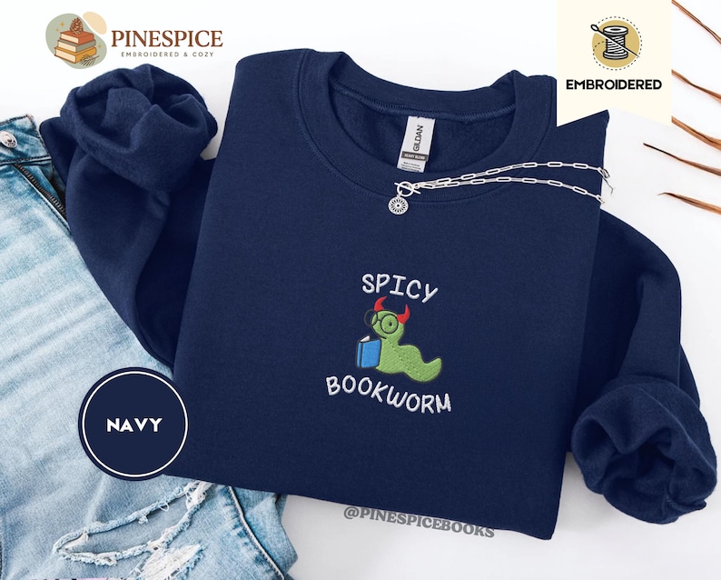 Puede incluir: Sudadera azul marino con el texto bordado "SPICY BOOKWORM" y un dise&ntilde;o de gusano de libro de dibujos animados. El dise&ntilde;o presenta un gusano verde con gafas y sosteniendo un libro. Un collar plateado est&aacute; drapeado sobre la sudadera.
