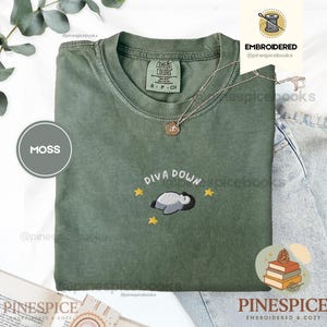 May include: A moss green t-shirt featuring an embroidered graphic of a sleeping penguin with the text "DIVA DOWN" and three yellow stars. The shirt has a crew neckline and a Comfort Colors brand label on the inside collar, paired with a thin gold necklace.