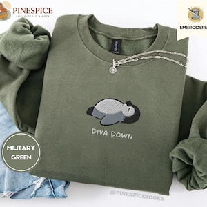 May include: Military green sweatshirt with an embroidered design of a sleeping creature and the words "DIVA DOWN". A silver necklace with a small pendant is on the sweatshirt. The brand name "PINESPICE" is visible.
