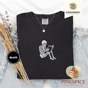 May include: Black Comfort Colors sweatshirt with a white embroidered skeleton reading a book. The sweatshirt has a crew neck and long sleeves. A silver necklace with a small pendant is visible. The word "BLACK" is in a black circle on the left.