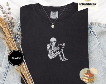 Puede incluir: Sudadera negra Comfort Colors con un esqueleto blanco bordado leyendo un libro. La sudadera tiene cuello redondo y mangas largas. Se ve un collar plateado con un pequeño colgante. La palabra "BLACK" está en un círculo negro a la izquierda.