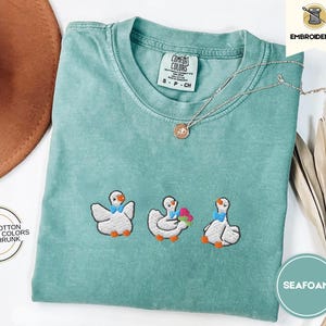 Peut inclure: T-shirt vert mousse avec des oies brodées. Les oies sont blanches avec des nœuds bleus et une oie tient un cœur rouge. Le t-shirt est 100% coton, couleurs confortables et pré-rétréci.
