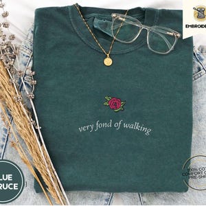 Puede incluir: Una camiseta de color verde azulado con la frase bordada "very fond of walking" y un dise&ntilde;o de rosa. Un collar dorado con colgante y gafas de montura transparente descansan sobre la camisa. La camisa est&aacute; doblada sobre una superficie de mezclilla.