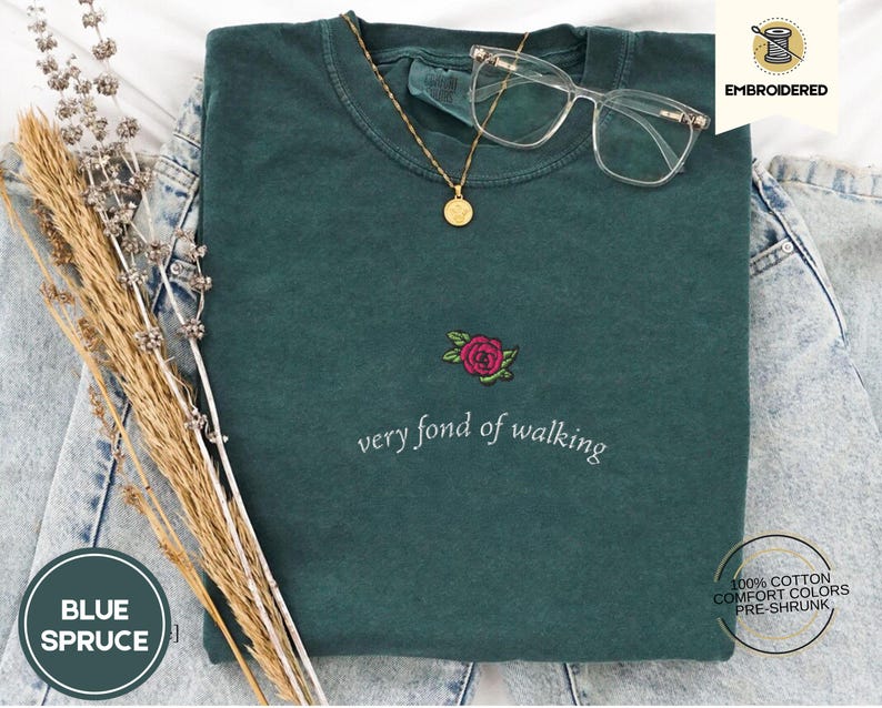 Puede incluir: Una camiseta de color verde azulado con la frase bordada "very fond of walking" y un dise&ntilde;o de rosa. Un collar dorado con colgante y gafas de montura transparente descansan sobre la camisa. La camisa est&aacute; doblada sobre una superficie de mezclilla.