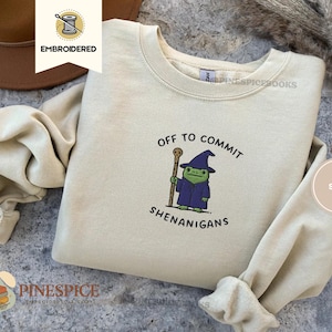 May include: Sand-coloured sweatshirt featuring the embroidered text "Off to Commit Shenanigans" and a cartoon frog wizard. The frog is wearing a purple robe and hat, holding a staff. The sweatshirt has a crew neck.