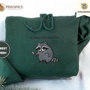 May include: Forest green hoodie with a drawstring and embroidered raccoon graphic holding a knife. The text "I require snacks" is above the raccoon. The hoodie is made of a soft material.