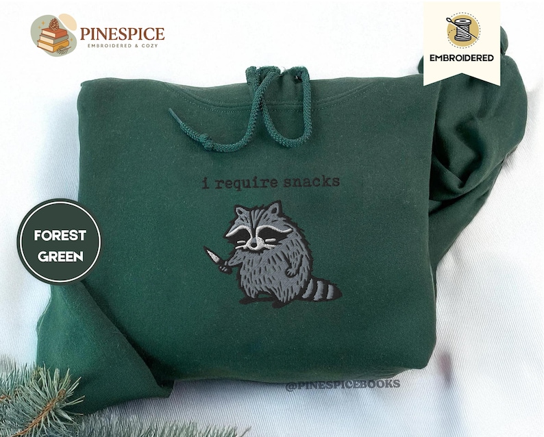 May include: Forest green hoodie with a drawstring and embroidered raccoon graphic holding a knife. The text "I require snacks" is above the raccoon. The hoodie is made of a soft material.
