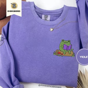 May include: Violet crewneck sweatshirt with an embroidered design of a green frog reading a purple book while sitting on a log. The sweatshirt has a relaxed fit and is made of 100% cotton. The text "VIOLET" is in a circle on the bottom right.