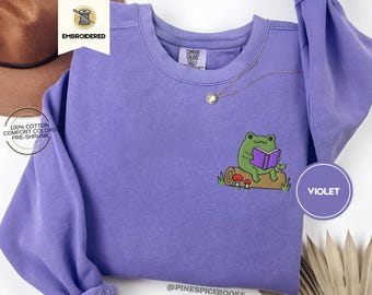 Può includere: Felpa girocollo viola con un disegno ricamato di una rana verde che legge un libro viola, seduta su un tronco. La felpa è in cotone 100%. La parola "VIOLET" è in un cerchio in basso a destra.