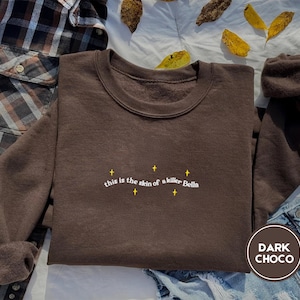 May include: A dark brown sweatshirt with the text "This is the skin of a killer Bella" in white lettering. The sweatshirt has a crew neck and long sleeves.
