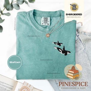Könnte beinhalten: Ein kurzärmeliges T-Shirt in Meerschaumgrün mit einer Stickerei von zwei schwarz-weißen Orcas auf der Brust. Das Shirt hat einen Rundhalsausschnitt und ein Comfort Colors Markenetikett am Innenkragen. Eine silberfarbene Halskette liegt auf dem Stoff.