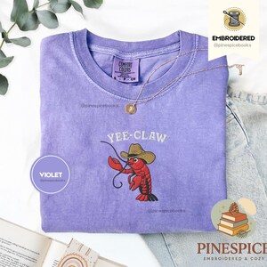 May include: A violet Comfort Colors t-shirt with an embroidered design. The design features a red lobster wearing a cowboy hat with the text "Yee-Claw". A gold necklace is draped across the shirt.