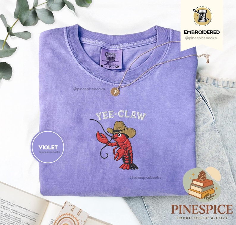 May include: A violet Comfort Colors t-shirt with an embroidered design. The design features a red lobster wearing a cowboy hat with the text "Yee-Claw". A gold necklace is draped across the shirt.