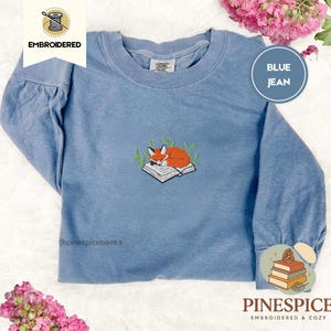 Puede incluir: Camiseta azul vaquera de manga larga con un diseño bordado de un zorro naranja durmiendo sobre un libro. La camiseta tiene cuello redondo y el texto "EMBROIDERED" y "BLUE JEAN". La marca "PINESPICE" también es visible.