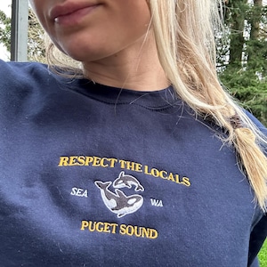 May include: A navy blue sweatshirt with the text "RESPECT THE LOCALS" embroidered in yellow. The text "SEA" and "WA" are also embroidered in yellow, along with two orcas in white. The text "PUGET SOUND" is embroidered in yellow below the orcas.