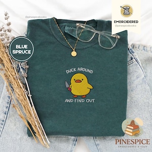 Puede incluir: Una camiseta azul abeto con un pato amarillo bordado sosteniendo un cuchillo, con el texto "DUCK AROUND AND FIND OUT". La camiseta también tiene un collar dorado y gafas transparentes. Parte de la colección Embroidered & Cozy.