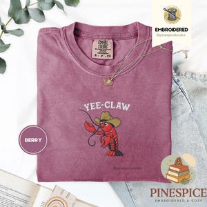May include: Berry-colored t-shirt with the embroidered phrase "Yee-Claw" and a cartoon crawfish wearing a cowboy hat. The shirt has a crew neck and short sleeves. The brand name "Comfort Colors" is visible on the tag.