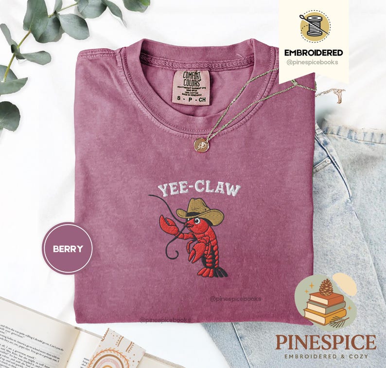 May include: Berry-colored t-shirt with the embroidered phrase "Yee-Claw" and a cartoon crawfish wearing a cowboy hat. The shirt has a crew neck and short sleeves. The brand name "Comfort Colors" is visible on the tag.