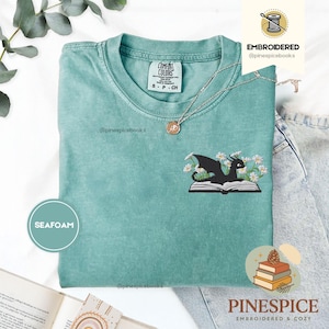 May include: Seafoam green t-shirt featuring an embroidered black dragon on an open book, encircled by white daisies. The shirt has a crew neck and short sleeves. A silver necklace with a pendant is visible.