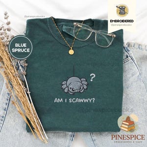 May include: Blue spruce t-shirt with an embroidered spider design and the text "AM I SCAWWY?". The shirt also features a gold necklace and clear-framed glasses. The shirt is displayed on a pair of light-wash jeans.