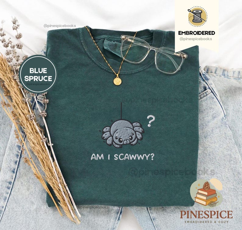 May include: Blue spruce t-shirt with an embroidered spider design and the text "AM I SCAWWY?". The shirt also features a gold necklace and clear-framed glasses. The shirt is displayed on a pair of light-wash jeans.