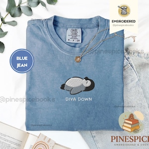 May include: A blue jean coloured t-shirt with an embroidered design of a sleeping cartoon character. The text "DIVA DOWN" is embroidered below the design. The shirt has a crew neck and short sleeves. The brand name "COMFORT COLORS" is visible on the shirt.