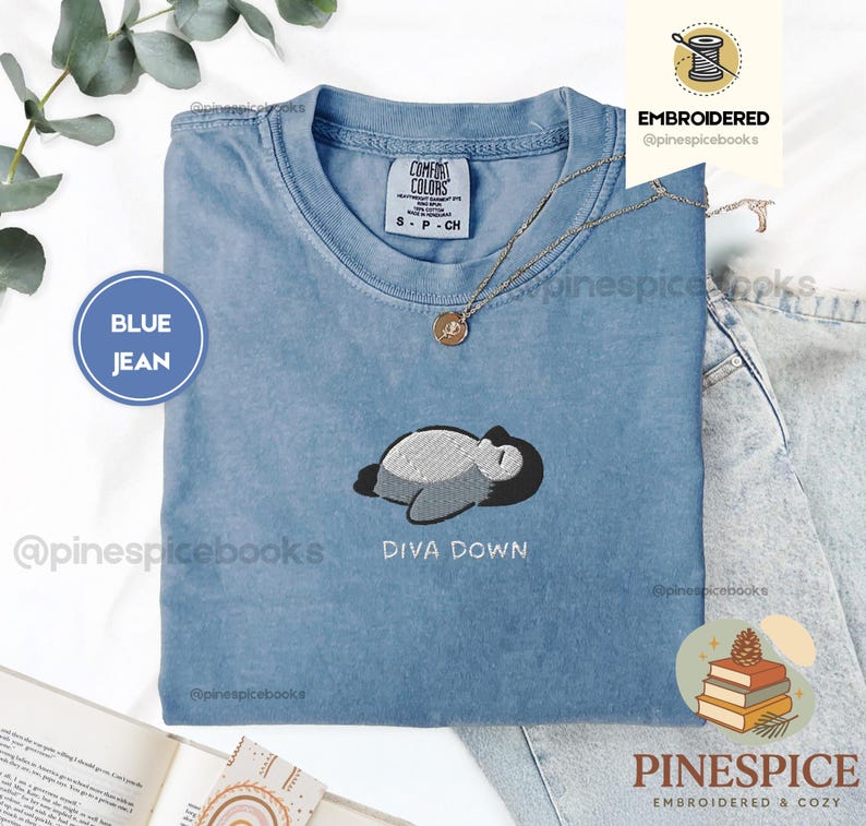 May include: A blue jean coloured t-shirt with an embroidered design of a sleeping cartoon character. The text "DIVA DOWN" is embroidered below the design. The shirt has a crew neck and short sleeves. The brand name "COMFORT COLORS" is visible on the shirt.