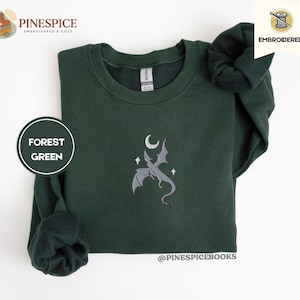 Puede incluir: Sudadera verde bosque con un diseño bordado de dragón, luna y estrellas. La sudadera de manga larga y cuello redondo tiene una etiqueta de marca. El texto "@pinespicebooks" es visible. Una prenda bordada y cómoda.