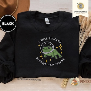 Può includere: Una felpa nera a maniche lunghe con il ricamo di una rana verde che indossa un casco da astronauta bianco. Il testo "I WILL SUCCEED BECAUSE I AM INSANE" è ricamato in bianco attorno alla rana, con piccole stelle gialle decorative.