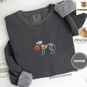 May include: A dark gray sweatshirt with a skeleton dog embroidered on the front, carrying an orange pumpkin-shaped candy bucket. The sweatshirt has the text "Pepper" on a circular label. The brand name "Pinespice" is also visible.