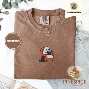 Puede incluir: Camiseta color espresso con un diseño bordado de un fantasma leyendo un libro en una silla naranja, con un gato negro. La camiseta tiene cuello redondo y mangas cortas. La marca "Comfort Colors" es visible en la camiseta.
