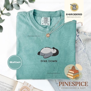 May include: Seafoam green t-shirt with an embroidered penguin design and the text "DIVA DOWN." The shirt is a Comfort Colors brand, size S-CH. A silver necklace with a pendant is visible. The shirt is displayed on a light background.