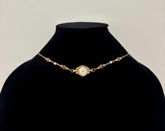 May include: A gold-coloured necklace with a small watch as its focal point. The watch has a cream-coloured face with black numerals and hands. The necklace is decorated with small red and white gemstones. The necklace is displayed on a black mannequin.