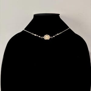 Puede incluir: Un collar plateado con un pequeño colgante de reloj cuadrado con una esfera color crema. El collar tiene delicadas cadenas y está adornado con pequeños adornos redondos. El colgante del reloj es el punto focal, mostrando una estética vintage.