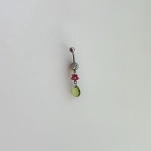 May include: A silver belly button ring with a dangling design. The top ball and center gem are clear, with a small red flower and a pear-shaped green gem. The jewellery is set against a white background.